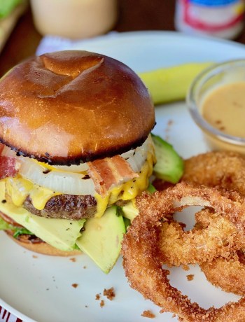 Juicy hamburger patty with a brioche bun, pickle, and copycat chick fil a sauce on a white plate and a side of homemade onion rings.